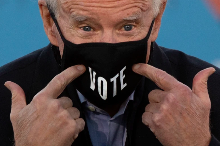 El presidente electo Joe Biden señala su máscara facial con la palabra "Vote" impresa en ella.